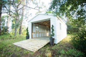 A white wooden shed with a ramp, ideal for storage or workshop, situated in a lush outdoor setting, perfect for garden or backyard use.