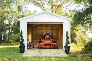 White wooden garage with lawn mower and small vehicle inside, surrounded by greenery and potted plants, ideal for storing outdoor equipment.